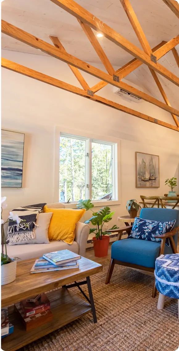 A cozy living space with exposed wooden beams, vibrant blue and yellow accent chairs, nautical-themed artwork, and natural lighting from a nearby window.