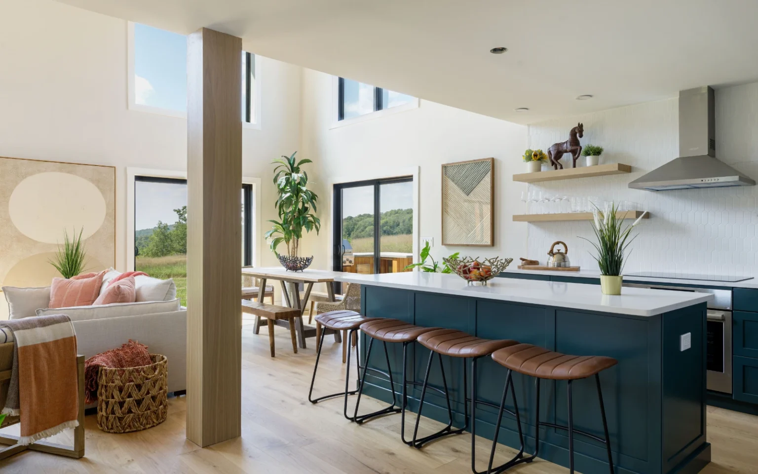 A bright, open-concept kitchen and living area with a blue island, brown leather barstools, large windows providing scenic outdoor views, and a mix of modern and natural decor elements.