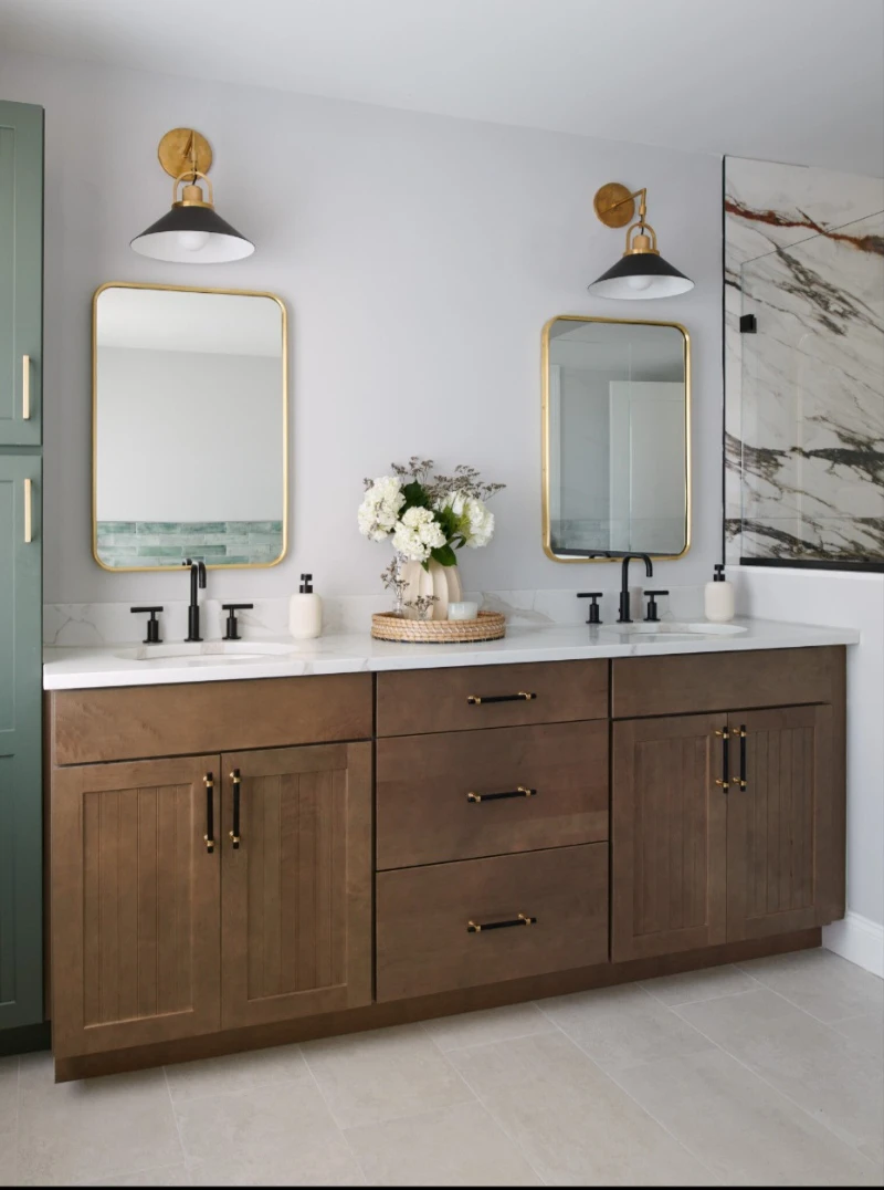 Elegant double-sink bathroom vanity with dark wood cabinetry, gold-accented mirrors, and black faucets, complemented by decorative greenery and neutral decor